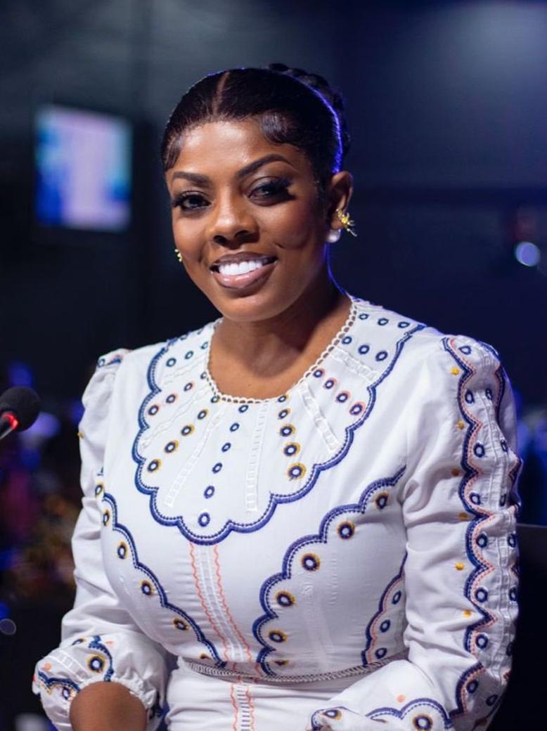 Nana Aba Anamoah smiling in a white embroidered dress in a broadcast studio